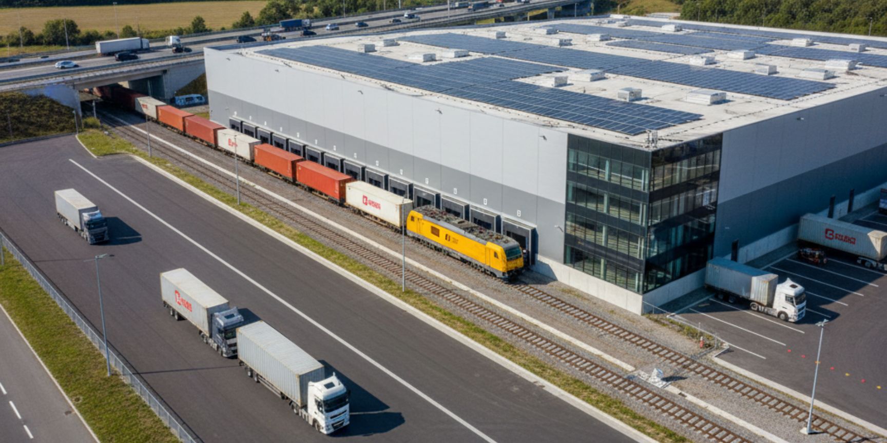 Eine Drohnenaufnahme zeigt eine große, moderne Logistikhalle mit Solarpaneelen auf dem Dach und einem direkten Gleisanschluss, an dem ein Güterzug mit Containern steht. Daneben befinden sich mehrere LKW auf breiten Zufahrtsstraßen, die auf eine nahegelegene Autobahn führen. Das Bild symbolisiert die effiziente Schnittstelle zwischen Schienen- und Straßentransport im bimodalem Verkehr und die Bedeutung von Logistikimmobilien als zentrale Umschlagpunkte.